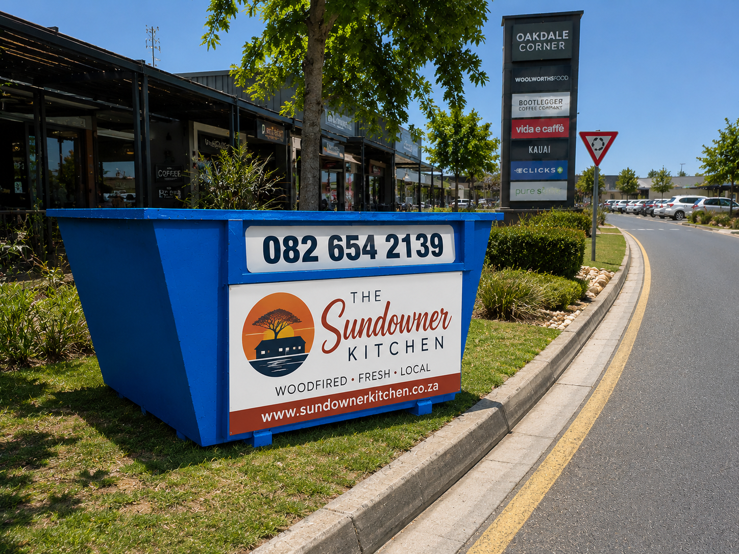 Blue skip carrying Sundowner Kitchen advertising near a local retail area.